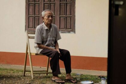 Elderly man sitting pensively on a wooden chair outside his home, reflecting solitude.