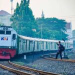 Passenger train arriving at a station with a man signaling next to the tracks amidst urban surroundings.