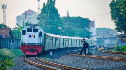 Passenger train arriving at a station with a man signaling next to the tracks amidst urban surroundings.